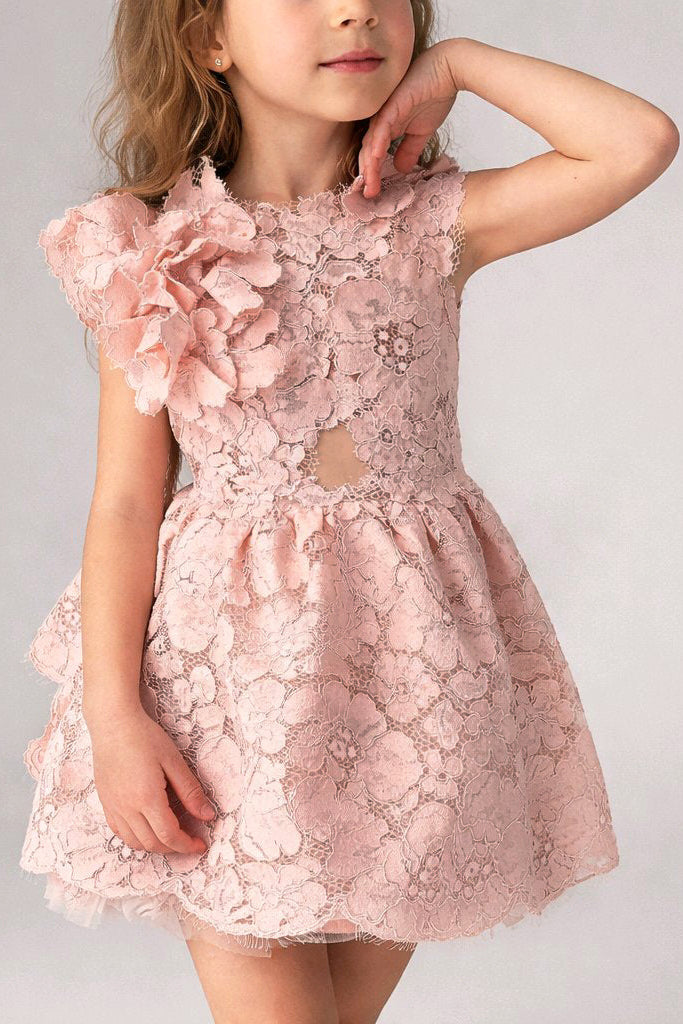 A young girl wearing a dusty pink floral lace dress with 3D ruffle details on the sleeves and a sheer lace overlay.