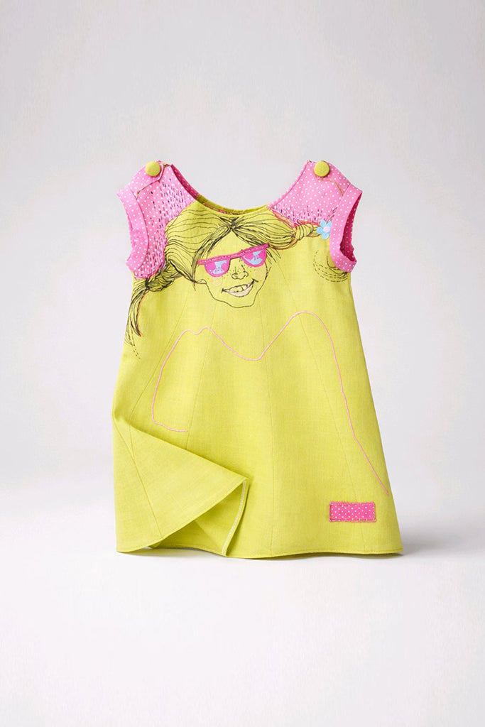 A children's sleeveless dress in bright yellow-green with pink crochet shoulder panels, a cartoon face graphic wearing pink sunglasses, and a small pink patch near the hem.