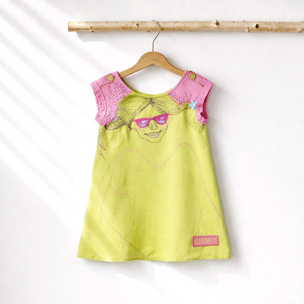 A sleeveless children's dress hanging on a wooden hanger against a white wall. The dress is lime green with a pink crocheted yoke and a whimsical embroidered face wearing pink sunglasses on the front.