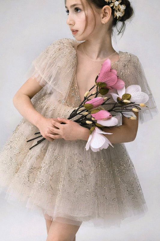 A young girl wearing a sparkly champagne tulle flower girl dress with sheer flutter sleeves, holding a bouquet of pink and white artificial flowers.