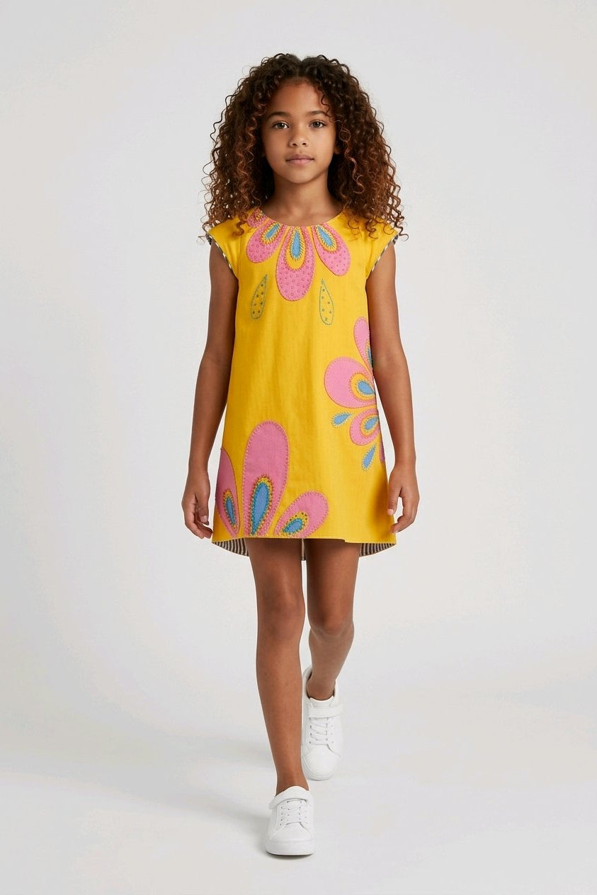 A young girl wearing a bright yellow sleeveless dress with pink and teal paisley patterns.
