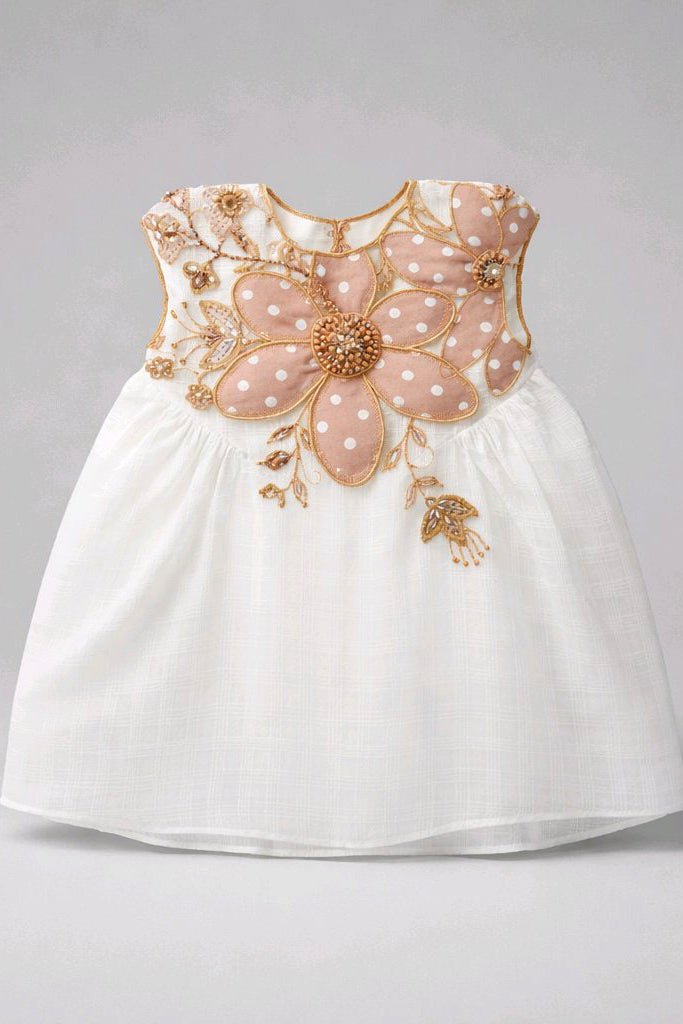 A sleeveless white baby dress featuring a large, decorative pink flower applique with gold trim and white polka dots on the bodice.