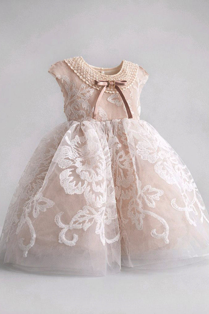 A toddler girl's formal party dress in a soft blush nude tulle with white floral lace overlays, featuring a crew neck with pearl-like embroidery, a brown satin bow, and cap sleeves.