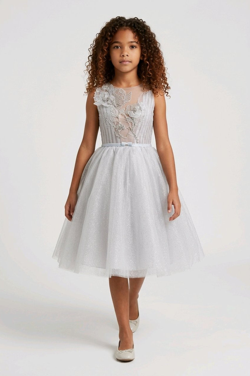 A young girl wearing a sleeveless white tulle dress with a lace bodice and a matching waist tie.
