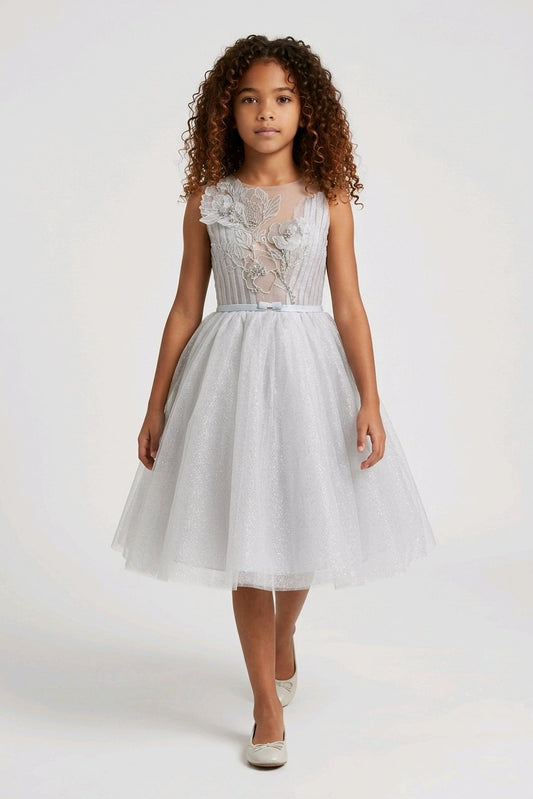 A young girl wearing a sleeveless white tulle dress with a lace bodice and a matching waist tie.